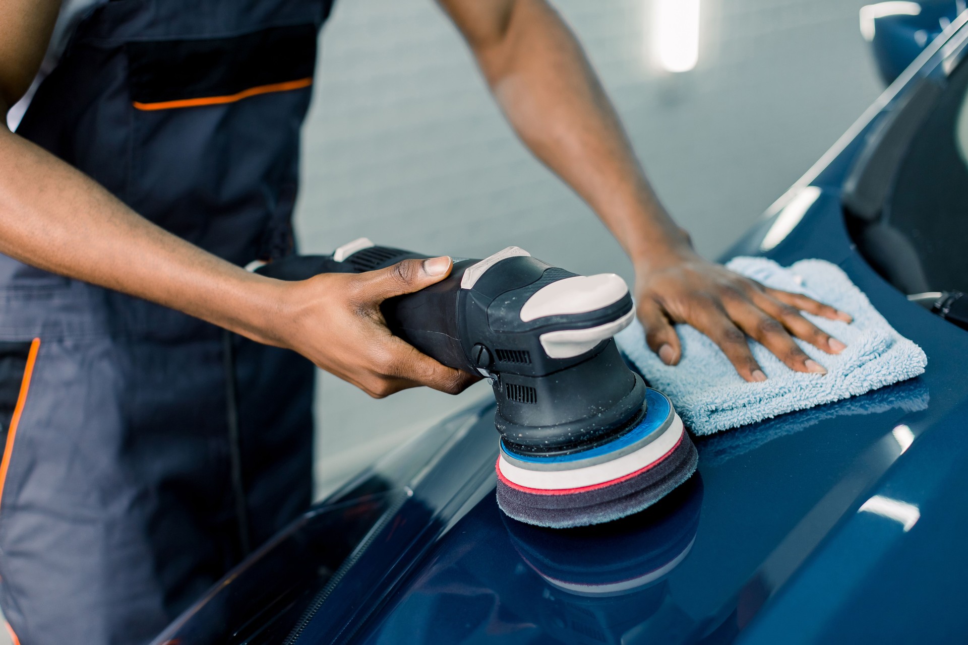 Car detailing and polishing concept. Hands of professional African man worker, with orbital polisher, polishing blue luxury car hood in auto repair shop